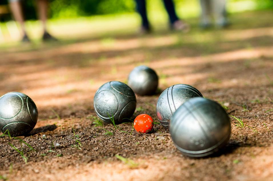 Gleise mit silbernen Boulekugeln und einer roten Kugel auf einem erdigen Boden in einem Park.